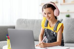 Studying At Home. Smiling pupil in pink headset sitting on couch at table using pc, writing essay or test exam, taking notes in textbook listening to teacher looking at screen during online lesson photo