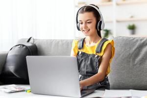 E-learning concept. Smiling teenager in headset studying online on laptop computer at home. Happy youth using PC to make home assignment, typing on keyboard, chatting with friends or doing homework photo