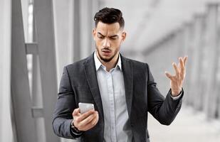 Portrait Of Worried Arab Businessman Reading Message On Smartphone While Standing In Airport Terminal, Anxious Middle Eastern Man Looking At Cellphone Screen, Emotionally Reacting To Bad News photo