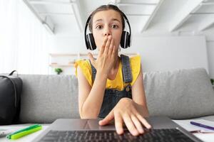 Portrait of young surprised girl in headset looking at screen with open mouth in amazement, reading news having online call on laptop covering mouth typing on keayboard, web camera point of view photo
