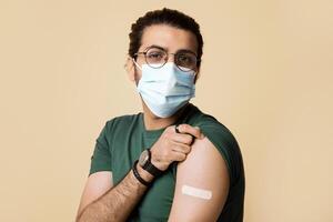 Healthy indian guy in face mask showing medical band on shoulder after vaccination against COVID-19, posing on beige studio background, closeup, copy space. Mass immunization concept photo