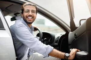 Excited indian guy in casual getting into auto, making test-drive before purchasing, visiting open-air car saloon, smiling at camera while sitting in nice brand new automobile, copy space photo