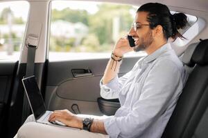 Positive young arab man freelancer working online while sitting at backseat in taxi, typing on laptop with blank screen and having phone conversation, mockup, looking at copy space, side view photo