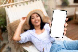 Happy Asian woman lying in hammock near RV, showing smartphone with blank screen on campsite, mockup for mobile app or website design. Selective focus. Cool summer vacation offer concept photo