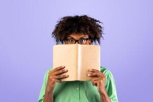 University exams. Shocked african american guy in glasses covering face with book, hiding behind it with eyes wide open, standing over blue studio background photo