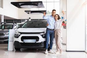 Joyful middle-eastern couple standing next to luxury white car and hugging at dealership automobile salon, rasising hands up, showing key from their new auto, cheerfully smiling at camera, full length photo