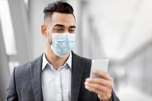 Arab Man In Medical Mask Using Smartphone While Waiting In Airport Terminal, Middle Eastern Businessman Checking Digital Vaccine Health Passport Certificate On Mobile Phone Or Browsing Internet photo
