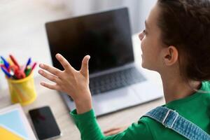 Online Call And Communication. Over shoulder view of teenage lady sitting at table, using laptop with black empty screen for virtual conference, talking waving hand to webcam, selective focus photo
