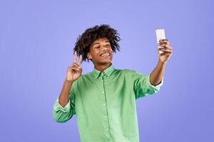 Excited african american guy taking selfie on smartphone and showing v-sign, having online chat or streaming live on web, standing over blue studio background photo