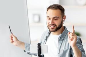 Happy male teacher giving online private lesson from home, pointing at empty blackboard with empty space. Millennial tutor communicating with pupils on webcam, using cellphone photo