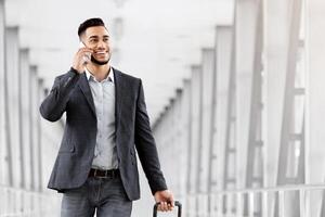 Handsome Arab Man Walking With Suitcase In Airport And Talking On Cellphone, Young Middle Eastern Businessman Booking Transfer By Phone After Arrival, Enjoying Pleasant Conversation, Copy Space photo