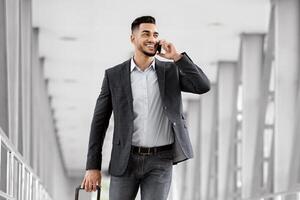 Handsome young arab businessman talking on cellphone and carrying suitcase in airport, smiling middle eastern man walking with luggage to flight gate and enjoying mobile conversation, copy space photo