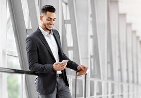 Handsome Arab Man Relaxing With Smartphone While Waiting For Flight In Airport, Smiling Middle Eastern Male Traveller Using Mobile Phone In Terminal, Booking Hotel Online Or Browsing Internet photo