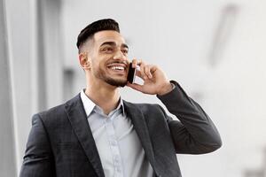 Business Trip. Smiling Arab Male Entrepreneur Holding Digital Tablet And Talking On Cellphone While Walking At Airport Terminal, Middle Eastern Businessman Enjoying Mobile Conversation, Copy Space photo