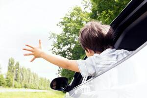 Rear view of little boy leaning out of car window and trying to catch wind with outstretched hand, small male child enjoying summer weather and automobile ride, on the move shot, closeup photo