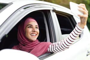 Joyful Muslim Woman In Hijab Taking Selfie While Sitting In Car, Happy Middle Eastern Female Leaning Out Hand And Making Photo With Smartphone During Ride, Enjoying Automobile Journey, Closeup