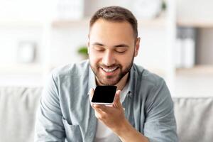 Cheerful millennial guy sitting on couch with cellphone at home, mockup for mobile app or website on screen. Young Caucasian man using modern gadget indoors. Contemporary technologies concept photo
