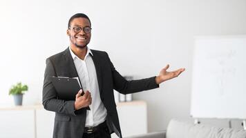 Friendly young African American psychologist with clipboard showing something with his hand at mental health clinic, copy space. Black male psychotherapist posing and smiling at office photo