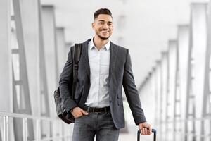 Portrait Of Handsome Happy Arab Man Walking In Airport Terminal With Suitcase, Smiling Young Middle Eastern Guy In Casual Clothes Going To Boarding Gate, Enjoying Upcoming Air Trip, Copy Space photo