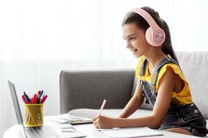 Online Lecture. Portrait of girl in pink headset sitting on couch at table using laptop looking at screen, writing essay or making test, taking notes in textbook, listening to teacher, free copy space photo