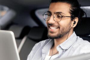 Smiling arab guy sitting at car back seat, listening to music while going somewhere by transportation service, using earphones and mobile application on digital tablet, copy space, closeup photo