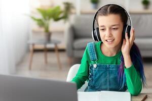 Good Connection, E-learning Concept. Portrait of teenafe girl touching wireless headphones, sitting at table, using pc computer and watching online tutorial, lecture or class, studying online at home photo