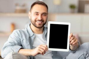Happy young man showing tablet computer with mockup for website or application on screen, indoors. Smiling millennial guy holding touch pad with copy space, selective focus photo