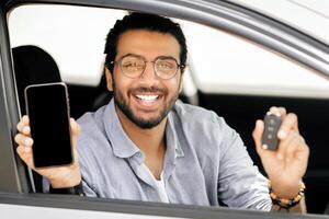 Cheerful middle-eastern guy sitting inside nice auto, showing car key and smartphone with blank screen, positive arab man recommending online service for car renting, moclup, copy space photo