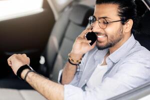 Happy young indian man in casual outfit sitting at car back seat, having phone conversation, checking time, going to airport by taxi, middle-eastern guy taking cab, copy space photo