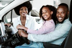 Happy black family buying new car, test driving automobile, smiling at camera in auto dealership. Afro parents and their daughter selecting luxury vehicle at showroom store photo