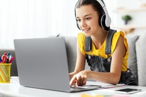 Kids And Technology Concept. Portrait of smiling girl sitting at table and using laptop computer, wearing wireless headphones, watching online course or movie, playing online game, free copy space photo