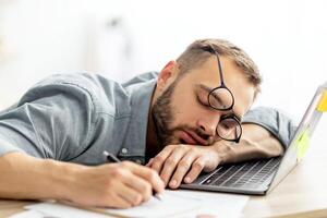 Exhausted millennial man sleeping on his office desk, next to laptop and documents, tired of overworking. Young male workaholic suffering from chronic fatigue at workplace photo
