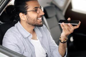 Cheerful middle-eastrn man recording audio message while taking taxi to office, sitting at car back seat and using mobile phone, sending girlfriend or business partner voice message, copy space photo