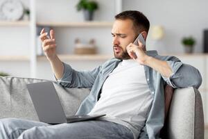 Stressed young businessman using laptop and making call on mobile phone from home office. Millennial Caucasian guy solving problem, overworking to meet project deadline, feeling upset photo