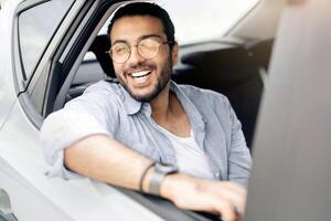 Cheerful arab guy passenger sitting at auto back seat, looking through window and smiling, enjoying car ride, closeup portrait. Happy indian man taking taxi, going to office, copy space photo