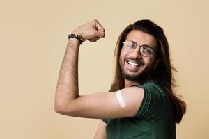 Handsome long-haired middle-eastern guy showing biceps with medical plaster on, got vaccinated against COVID-19 while pandemic, feeling strong and positive, beige studio background photo