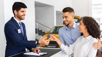 Happy middle-eastern couple buying car in huge showroom, taking automobile key from young handsome salesman, side view. cheerful arab man and woman purchasing beautiful auto, hugging and smiling photo