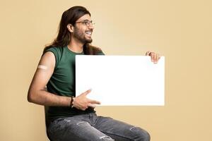 Cheerful middle-eastern vaccinated man with empty board posing on beige studio background, looking at copy space and smiling. Happy young arab guy recommending vaccination while COVID-19 pandemic photo
