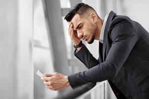 Worried middle eastern businessman looking at smartphone screen while waiting at airport terminal, anxious arab man touching forehead, reading unpleasant message or suffering business problems photo