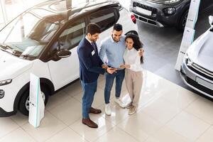 Top view of cheerful middle-eastern young couple having conversation with good-looking sales assistant at auto showroom, checking papers or car specification, full length photo