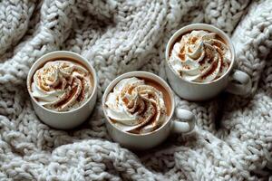 Three mugs of hot chocolate with whipped cream and cinnamon on a cozy knitted blanket photo