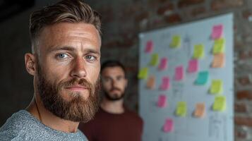 Confident businessman with beard looking at camera, colleague in background, brainstorming session photo