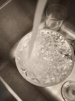 Crystal bowl filling with tap water in a stainless steel kitchen sink, with a drinking glass nearby, in prague, precious resource, take care of it World Water Day photo