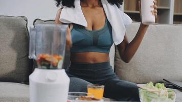 A woman in a sports bra top sits on a couch with a blender and a glass of juice video