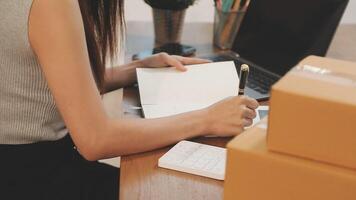 A woman is writing in a notebook while sitting at a desk with a laptop and a box of video