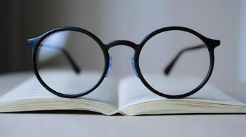 Black round glasses rest on closed book against pure white background. Minimalist composition emphasizes elegant simplicity through professional product photography with soft shadows and studio refi photo