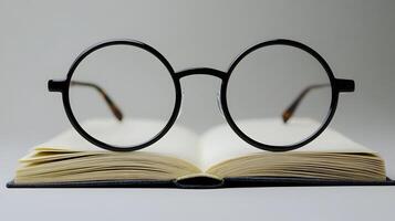 Black round glasses rest on closed book against pure white background. Minimalist composition emphasizes elegant simplicity through professional product photography with soft shadows and studio educ photo