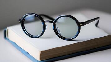 Black round glasses rest on closed book against pure white background. Minimalist composition emphasizes elegant simplicity through professional product photography with soft shadows and studio shad photo