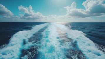 Ocean wake pattern stretches behind cruise ship in aerial view. Deep blue sea meets bright sky with white clouds, while turquoise foam trails create dynamic water movement on sunny day. blue photo