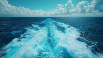 Ocean wake pattern stretches behind cruise ship in aerial view. Deep blue sea meets bright sky with white clouds, while turquoise foam trails create dynamic water movement on sunny day. oceanic photo